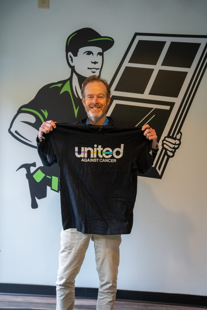 Smiling man holding a black "United Against Cancer" t-shirt