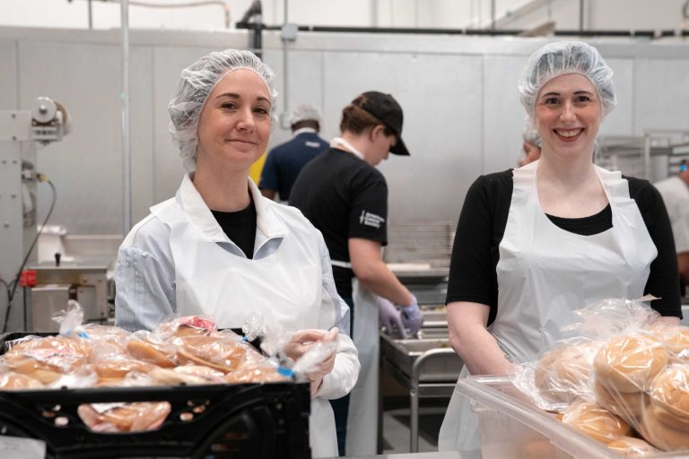 Two women wearing hairnets smiling next to hamburger buns